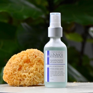 May include: A clear glass bottle of Lunasea Botanicals rejuvenating facial mist with snow mushroom. The bottle has a white cap and a label with the text "TREMELLA MOONSHINE". Beside the bottle is a yellow, sponge-like object. The background is a blurred green.
