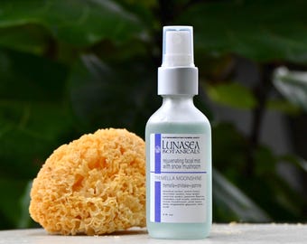 May include: A clear glass bottle of Lunasea Botanicals rejuvenating facial mist with snow mushroom. The bottle has a white cap and a label with the text "TREMELLA MOONSHINE". Beside the bottle is a yellow, sponge-like object. The background is a blurred green.