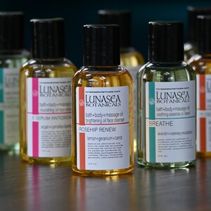 May include: Six amber glass bottles with black caps and labels. The labels are white with black text and feature the brand name "LUNASEA BOTANICALS". Each bottle has a different name and description of the product inside. The names are "THERAPY", "SERUM ANTIOXIDANT", "ROSEHIP RENEW", "BREATHE", and "WINTER RELAXER".