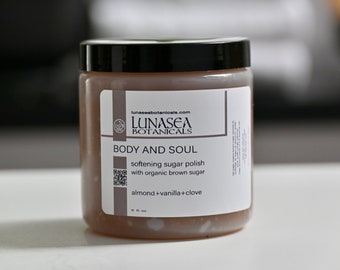 May include: A clear glass jar with a black lid containing a brown sugar scrub. The label reads "lunaseabotanicals.com LUNASEA BOTANICALS BODY AND SOUL softening sugar polish with organic brown sugar almond+vanilla+clove 8 fl. oz."