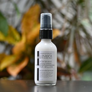 Op de afbeelding: Een witte fles met olievrije gezichts-serum met een zwarte pomp. De fles is gelabeld met "LUNASEA BOTANICALS" en "ACTIVE WILLOW". De fles staat op een donker oppervlak.