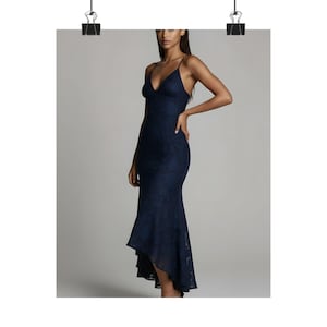 May include: A navy blue, floor-length dress with a V-neckline and spaghetti straps. The dress features a fitted silhouette that flares out at the hem, creating an asymmetrical, high-low design. The fabric has a subtle floral pattern.
