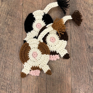 May include: Three handmade crochet cow-shaped coasters. Each coaster features a white base with black or brown spots, a pink center, and a tail. The coasters are designed to resemble a cow's rear.