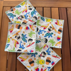 Pode incluir: Cinco bases para copos quadradas de tecido branco sujo com uma estampa tropical colorida com surfistas, golfinhos, sushi e palmeiras. Cada base para copos tem uma borda costurada verde claro e um botão no centro.