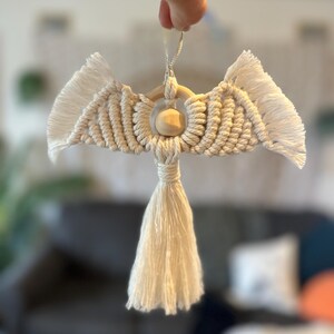 May include: A handmade macrame angel ornament with a wooden bead head and a long tassel. The wings are made of knotted cotton cord with fringed edges. The angel is suspended by a silver string.
