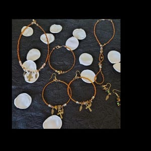 May include: An assortment of handmade jewelry, including necklaces, bracelets, and earrings. The pieces feature gold-colored beads and charms, with some adorned with feathers and cross pendants. The jewelry is arranged on a dark surface with white petal-shaped accents.