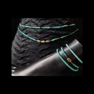 May include: A multi-strand beaded anklet featuring small turquoise and green glass seed beads. The design includes three small, oval-shaped gold-toned metallic beads centered on the strands, creating a layered appearance with a delicate, uniform texture.