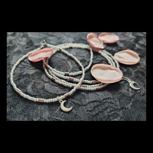 May include: A set of beaded necklaces or bracelets featuring small white and metallic copper-toned cylindrical beads arranged in a repeating pattern. Each strand is accented with a small silver-colored crescent moon charm and light pink petals.