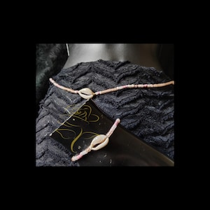May include: A beaded waist chain and a matching beaded bracelet, both featuring a single white cowrie shell as a central focal point. The jewelry is crafted with small, light pink and white beads arranged in a delicate, uniform pattern.