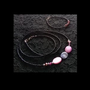 May include: A black beaded necklace and bracelet set. The necklace features pink and gray oval beads, with smaller purple and silver beads. The bracelet has brown beads. The jewelry is displayed on a black lace surface.