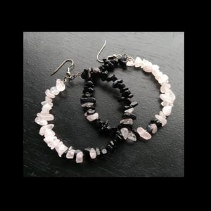 May include: Two pairs of hoop earrings. One pair features light pink, irregular-shaped stones, while the other has black stones. Both pairs have silver-colored ear wires and are displayed on a dark surface.