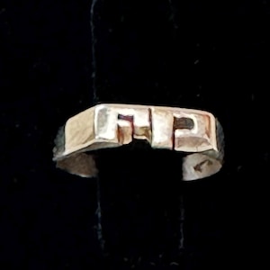 May include: A silver-toned ring with the letters "FP" in a blocky font. The ring is set against a black background, highlighting its geometric design. The ring's surface appears slightly textured, adding to its unique appeal.