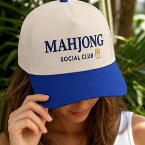 May include: A beige baseball cap with a blue brim and a blue button on top. The front features the dark blue text "MAHJONG SOCIAL CLUB" with a small gold-colored rectangular icon depicting a mahjong tile with a bamboo character design.