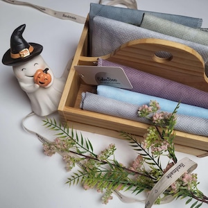 Op de afbeelding: Een houten mandje bevat gevouwen stukken stof in blauwe, paarse en grijze tinten. Een wit keramieken spookfiguurtje met een zwarte heksenhoed dat een kleine oranje pompoen vasthoudt, staat ernaast. Een lint met de tekst Handmade is zichtbaar.