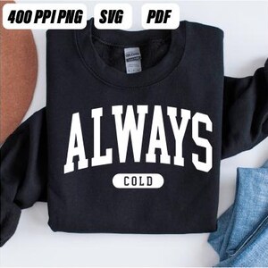 以下が含まれることがあります： 太字のアーチ型フォントで「ALWAYS COLD」と書かれた白い文字が特徴の黒いスウェットシャツ。「COLD」という単語は「ALWAYS」の下に、より小さく長方形のフォントで書かれています。画像には「400 PPI PNG SVG PDF」という文字も含まれています。