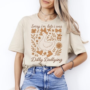Puede incluir: Camiseta beige con una ilustración caprichosa de un ganso con botas, rodeado de elementos florales y lazos. El texto dice "Sorry I'm late I was Dilly Dallying". Una camiseta informal y cómoda para el uso diario.