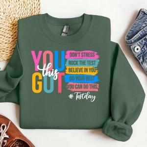 Może przedstawiać: Ciemnozielona bluza z napisem "YOU GOT this" w kolorowych blokowych literach. Bluza ma również frazy "DON'T STRESS, ROCK THE TEST, BELIEVE IN YOU, DO YOUR BEST, YOU CAN DO THIS" i hashtag "#Testday".