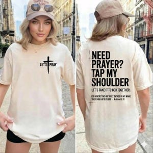 Puede incluir: Camiseta color crema con el texto "LET'S PRAY" y un gráfico de cruz en la parte delantera. La parte trasera de la camiseta presenta el texto "NEED PRAYER? TAP MY SHOULDER" y un versículo bíblico.