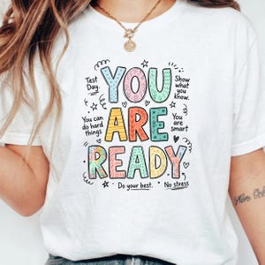 Puede incluir: Camiseta blanca con un gráfico colorido que dice "YOU ARE READY". El diseño incluye las frases "Test Day", "You can do hard things", "Show what you know" y "You are smart".