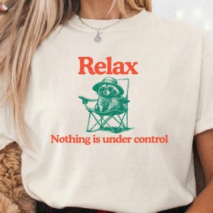 May include: Cream-colored t-shirt with the word "Relax" in red block letters above a green illustration of a raccoon in a beach chair. Below the illustration, the text reads "Nothing is under control."
