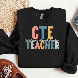 Könnte beinhalten: Schwarzes Sweatshirt mit der Aufschrift "CTE TEACHER" in bunten Blockbuchstaben. Die Worte "Career and Technical Education" sind in kleinerer, kursiver Schrift geschrieben. Das Sweatshirt ist gefaltet.