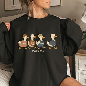 Puede incluir: Sudadera negra con un diseño caprichoso de cuatro patos, cada uno vestido con diferentes atuendos y etiquetado con palabras como "Shirley", "Good-ness" y "Mercy". El texto "Salmo 23:6" está impreso debajo. Un estilo casual y cómodo.