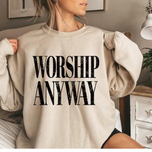 May include: A beige sweatshirt with the words "WORSHIP ANYWAY" printed in bold, black letters. The sweatshirt has long sleeves and a crew neck. The image is a close-up of the sweatshirt.
