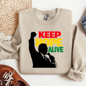 Puede incluir: Sudadera beige con las palabras "KEEP HOPE ALIVE" en rojo, amarillo y verde. Una silueta negra de una persona con el puño en alto y un micrófono aparece en la parte delantera. La sudadera está doblada.