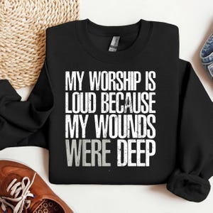 Peut inclure: Sweat-shirt noir avec l'inscription blanche "MY WORSHIP IS LOUD BECAUSE MY WOUNDS WERE DEEP". Le sweat-shirt est plié, avec un aperçu d'une chaussure marron et d'un jean en arrière-plan.