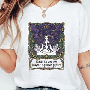 May include: White t-shirt featuring a graphic of a meditating figure with long purple hair, crystals, and mushrooms. The design includes the text "Maybe it's woo-woo. Maybe it's quantum physics."