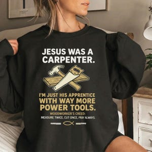 Puede incluir: Sudadera negra con el texto "JESUS WAS A CARPENTER." y "I'M JUST HIS APPRENTICE WITH WAY MORE POWER TOOLS." Un martillo y una sierra están cruzados sobre dos tablones de madera. El texto "WOODWORKER'S CREED: MEASURE TWICE, CUT ONCE, PRAY ALWAYS."