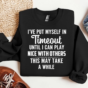 May include: Black sweatshirt with the text "I'VE PUT MYSELF IN Timeout UNTIL I CAN PLAY NICE WITH OTHERS THIS MAY TAKE A WHILE" in white. The sweatshirt is folded, with a glimpse of a brown shoe and a woven basket in the background.