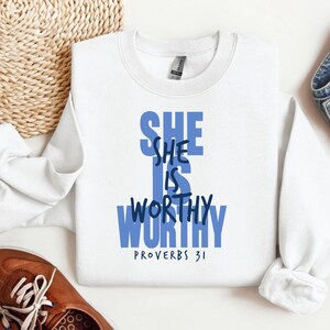 May include: A white sweatshirt with the text "SHE IS WORTHY" in blue, with "PROVERBS 31" below. The sweatshirt is on a white surface, with a woven basket and a brown shoe in the background.