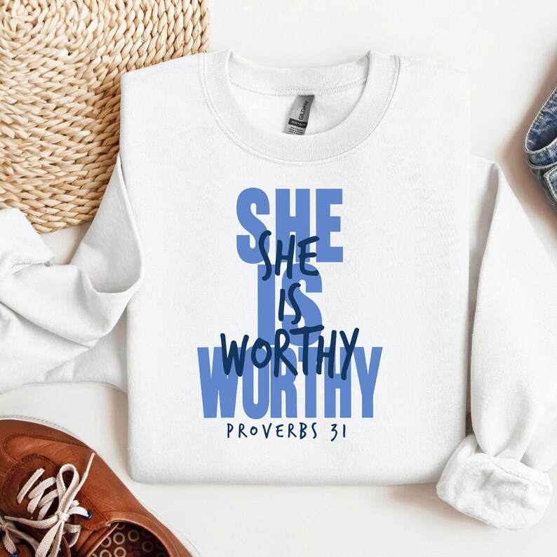 May include: A white sweatshirt with the text "SHE IS WORTHY" in blue, with "PROVERBS 31" below. The sweatshirt is on a white surface, with a woven basket and a brown shoe in the background.