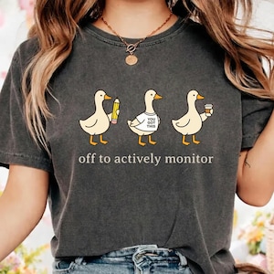 May include: Dark gray t-shirt featuring three cartoon ducks. One duck holds a pencil, another wears a shirt that says "You Got This," and the third holds a coffee cup. The text "off to actively monitor" is printed below the ducks.