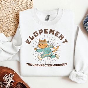 May include: White sweatshirt with the word "ELOPEMENT" in brown block letters above a cartoon cat wearing a blue tracksuit and headband. The cat is running, with the text "THE UNEXPECTED WORKOUT" below the cat.