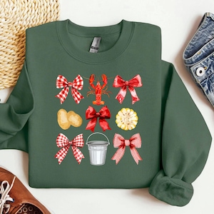 May include: Green sweatshirt with a design featuring a red lobster, red bows, corn, potatoes, and a metal bucket. The bows have a red and white checkered pattern. The design is centered on the front of the sweatshirt.