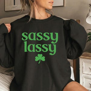 May include: Black sweatshirt with the words "sassy lassy" in green, along with a green shamrock. The sweatshirt is a crew neck style.