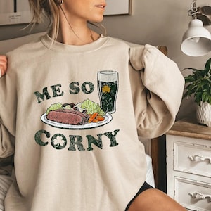 May include: Beige sweatshirt with the text "ME SO CORNY" in green, featuring a graphic of a plate with food and a pint of green beer. The graphic includes a shamrock. The sweatshirt has long sleeves.