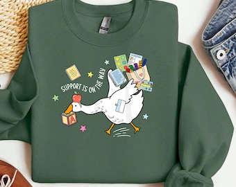 May include: Green sweatshirt with a white goose graphic carrying school supplies. The goose has an apple on its head and a block with the letter "A". The text "SUPPORT IS ON THE WAY" is printed on the sweatshirt.