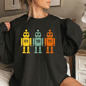 Peut inclure: Sweat-shirt noir avec trois illustrations de robots colorés. Les robots sont jaunes, turquoise et orange, chacun avec un numéro : 01, 10 et 11. Le design a une esthétique rétro.