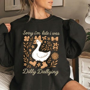 May include: Black sweatshirt with a white goose wearing boots and a bow, surrounded by floral and bow graphics. Text reads "Sorry I'm late I was Dilly Dallying". A casual, comfortable top.
