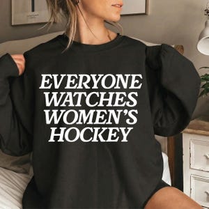 May include: Black sweatshirt with the text "EVERYONE WATCHES WOMEN'S HOCKEY" in white block letters. The sweatshirt has a crew neck and long sleeves. The image is a close-up of the sweatshirt.