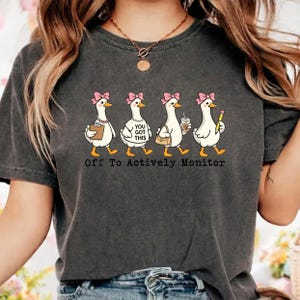 May include: Dark gray t-shirt featuring a line of cartoon geese with pink bows, each carrying different items. The geese are walking and the text "Off To Actively Monitor" is printed below them. The shirt is a casual style.