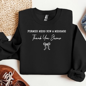 Könnte beinhalten: Schwarzes Sweatshirt mit weißem Text, der "FORMER MESS NOW A MESSAGE" und "Thank You Jesus" sowie eine Schleifengrafik zeigt. Das Sweatshirt liegt gefaltet auf einer hellen, gewebten Oberfläche.