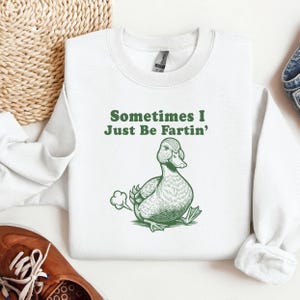 Puede incluir: Sudadera blanca con el texto "Sometimes I Just Be Fartin'" en verde. Una ilustración verde de un pato está debajo del texto, con una nube de gas saliendo de su parte trasera.