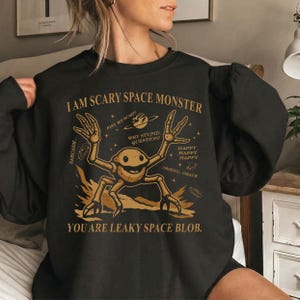 May include: Black sweatshirt with a humorous graphic. The design features a cartoon space monster with the text "I AM SCARY SPACE MONSTER" and "YOU ARE LEAKY SPACE BLOB." Additional text includes "FIST MY BUMP!" and "WHY STUPID, QUESTION?"