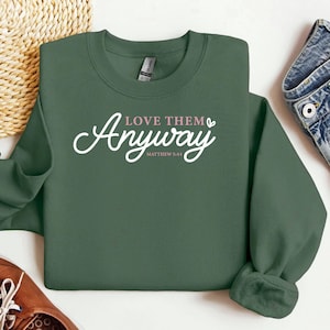 May include: A dark green sweatshirt with the words "Love Them Anyway" in white script and the Bible verse Matthew 5:44. The sweatshirt has a crew neck and long sleeves.