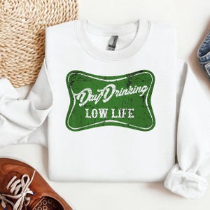 May include: White sweatshirt with a green, distressed graphic that reads "Day Drinking LOW LIFE". The graphic is in a retro style. The sweatshirt is made of a soft material.