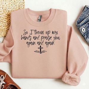 Op de afbeelding: Een lichtroze sweatshirt met de tekst "So I throw up my hands and praise you again and again" in zwart schrift. Onder de tekst staat een afbeelding van een open boek met een kruis. De sweatshirt heeft lange mouwen en een ronde hals.